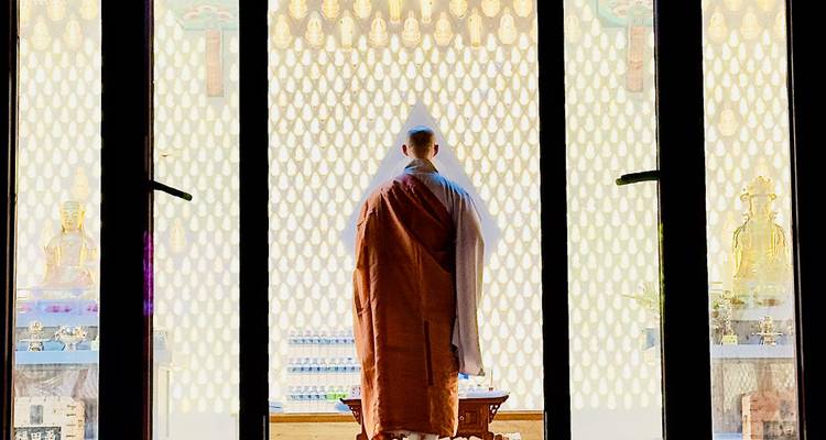 Moine en robes traditionnelles se tenant devant un mur à motifs illuminé à l'intérieur du temple, vu à travers l'encadrement de la porte
