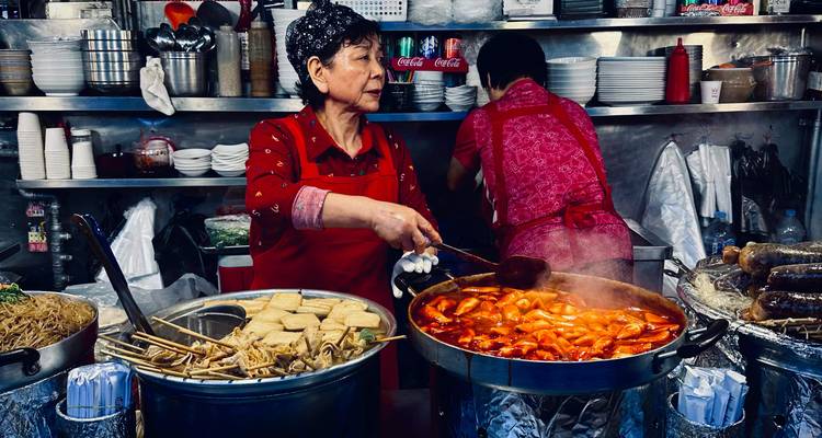 Étal de nourriture du marché coréen où une femme portant un tablier rouge prépare des tteokbokki et des brochettes dans de grandes casseroles