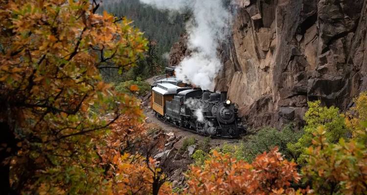 Un train à vapeur historique traverse péniblement un canyon rocheux étroit entouré d'un feuillage automnal coloré.