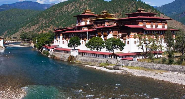 Grand monastère bhoutanais traditionnel au bord d'une rivière avec des montagnes en arrière-plan.