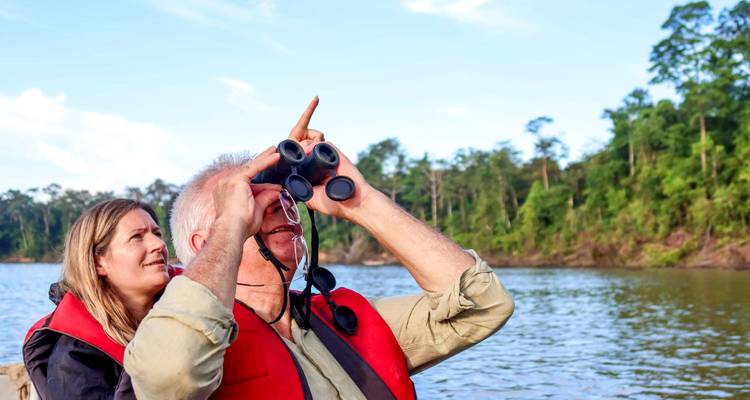 Mann mit Fernglas und Frau neben ihm beobachten den Dschungel von einem Boot aus auf einem breiten tropischen Fluss.