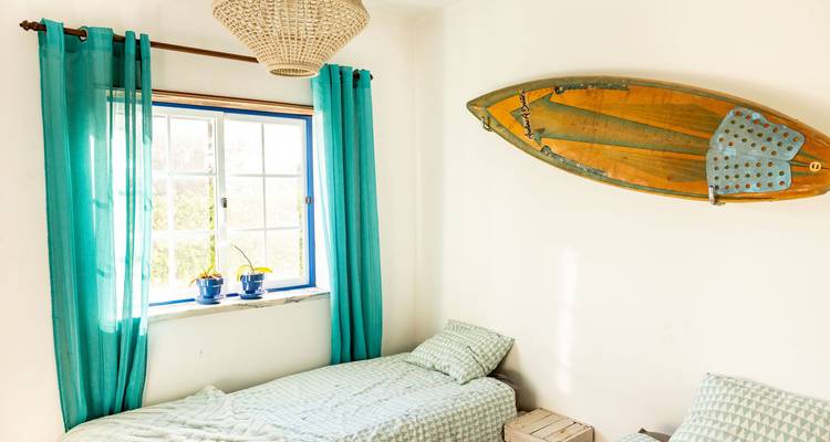 Intérieur de chambre avec un lit simple, une fenêtre avec des rideaux bleus, et une planche de surf au mur.