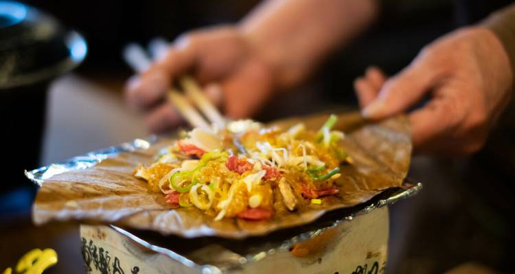 Hände servieren ein in Blätter gewickeltes warmes Gericht mit misogewürztem Gemüse auf einem kleinen Tischgrill.