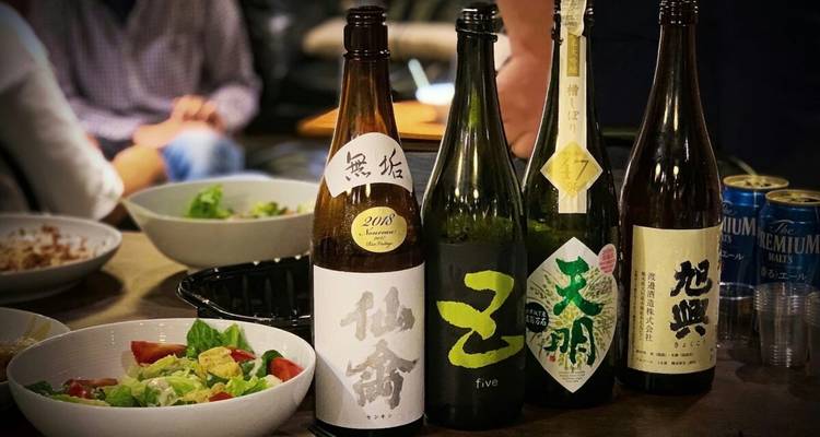 Sortiment von Sake-Flaschen, die auf einem Tisch während einer ungezwungenen Verkostung mit Snacks im Hintergrund angeordnet sind.