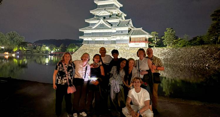 Eine Reisegruppe posiert fröhlich vor einem angestrahlten japanischen Schloss, das sich in dem umgebenden Burggraben bei Nacht spiegelt.