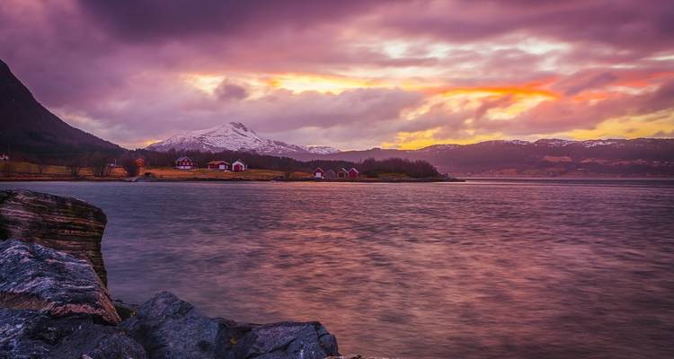 Coucher ou lever de soleil sur un fjord calme avec des maisons and des montagnes lointaines.