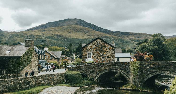 Historisches Dorf mit Steinbrücke und Hügeln im Hintergrund.