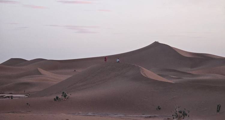 Twee mensen zitten bovenop een zachte zandduin bij zonsondergang, omringd door zacht verlichte woestijnlandschappen.