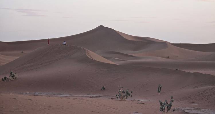 Avondaanzicht van gebeeldhouwde zandrichels met twee kleine figuren die op een top staan tegen een pastelkleurige hemel.