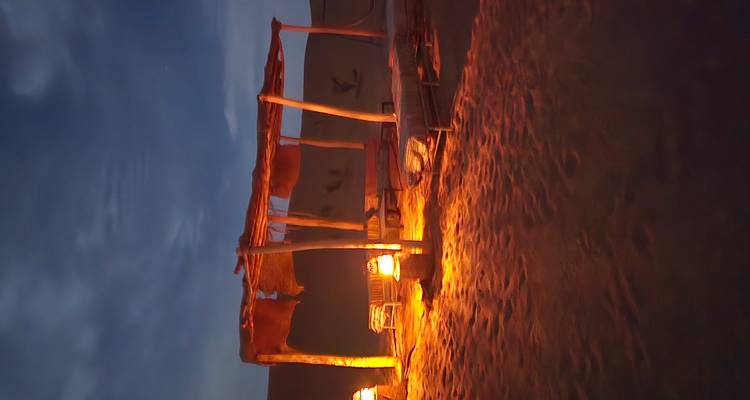 Nachtelijke woestijn lounge ruimte verlicht door lantaarns onder een gemoedvolle blauwe hemel, met kussens en lage tafels op het zand.