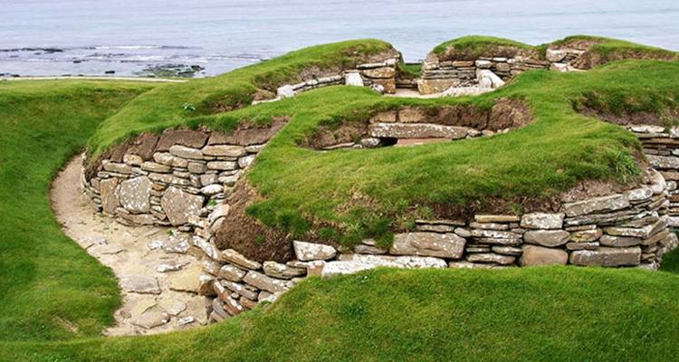 Ruines d'un ancien village de pierre recouvertes d'herbe surplombant la mer par une journée nuageuse.