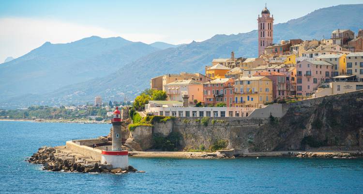 A scenic view of a coastal town with colorful buildings and a lighthouse, surrounded by mountains.