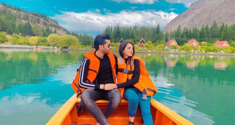 Un couple romantique en gilets de sauvetage assis dans une barque orange sur un lac alpin turquoise avec un chalet en arrière-plan.