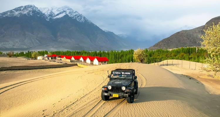 Jeep traversant les dunes dorées du désert de Shigar avec des huttes aux toits rouges et des pics enneigés en arrière-plan