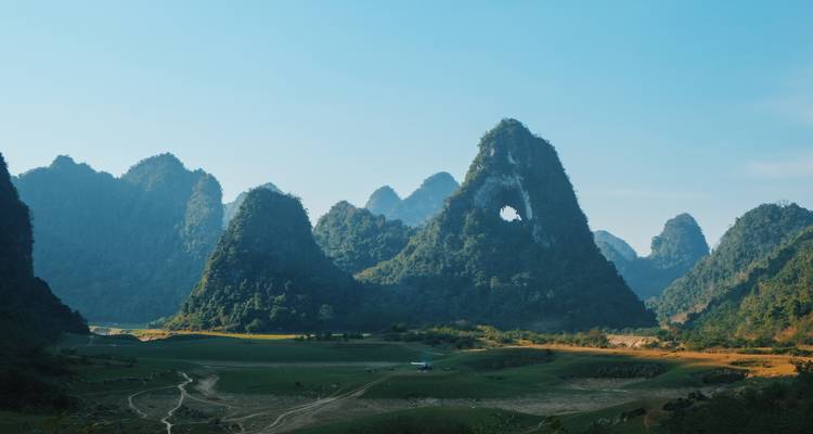 Dramatic karst mountains with a natural arch tower over a quiet green valley under a clear blue sky.