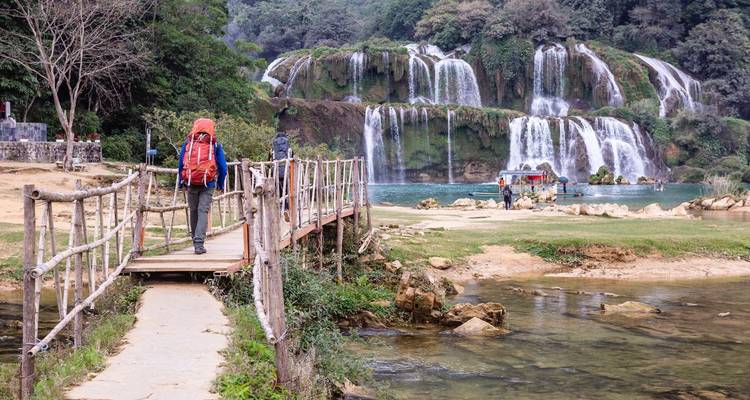 A backpacker with an orange pack crosses a rustic bridge toward a multi-tiered jungle waterfall.