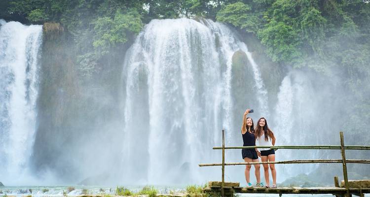 Two young women pose for a selfie on a wooden platform in front of a powerful misty waterfall.