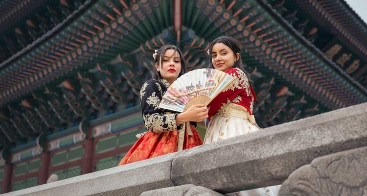 Two women in traditional attire holding a fan, in front of a historic building.