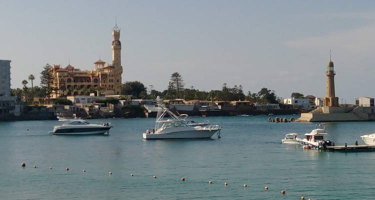 Vue sur le front de mer avec des bateaux, un grand bâtiment et un phare en arrière-plan.