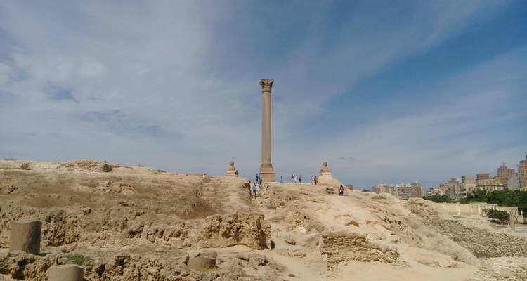 Ancienne colonne et statues de sphinx avec un arrière-plan de ville sous un ciel bleu.