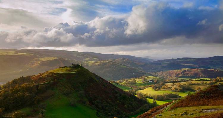 Vue panoramique d'un paysage vallonné sous un ciel nuageux avec des rayons de soleil qui percent.