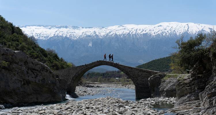 Trois personnes marchant sur un vieux pont de pierre au-dessus d'une rivière avec des montagnes enneigées en arrière-plan.