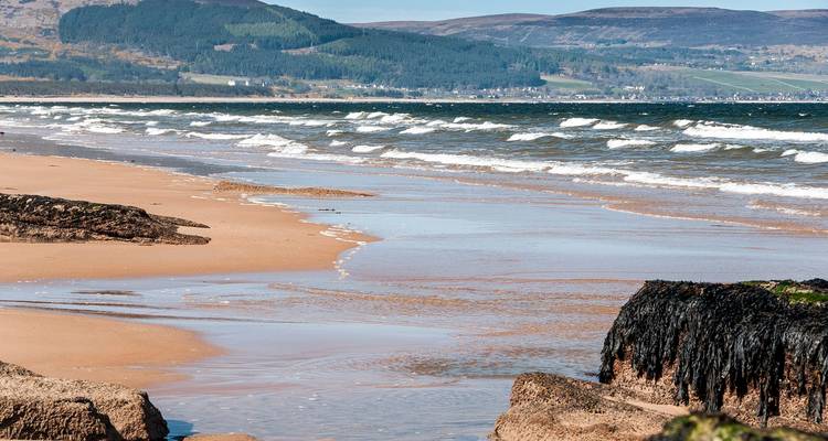 Plage de sable avec des vagues qui s'écrasent sur le rivage, collines en arrière-plan.