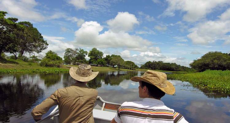 Two travellers wearing hats on a small boat cruising a reflective river in lush wetland