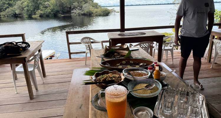 Configuration de repas en plein air sur une terrasse en bois avec vue sur l'eau, plusieurs plats et boissons sur la table.