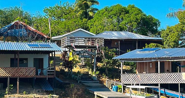 Hébergement simple au bord de la rivière avec plusieurs bâtiments rustiques, sous un ciel bleu dégagé.