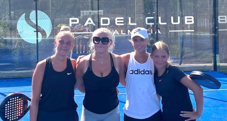 Cuatro mujeres sonriendo y posando en un club de pádel con raquetas en la mano.