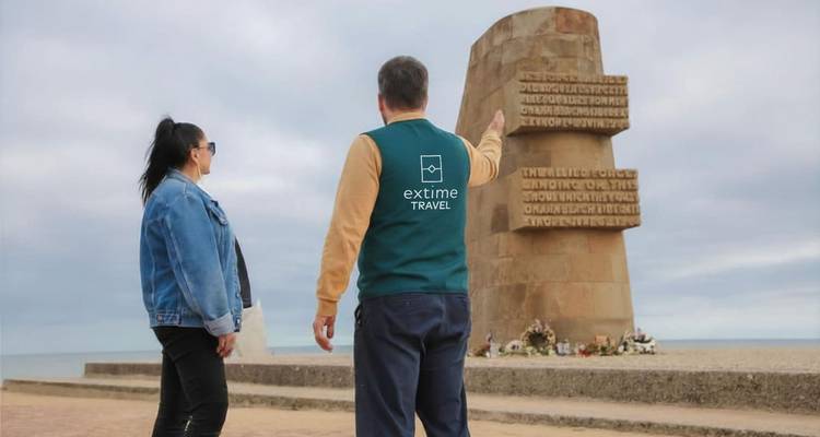Der Reiseführer deutet auf ein Denkmal am Meer hin, das an einem bewölkten Tag in der Normandie zu sehen ist.