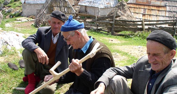 Tres ancianos sentados en la hierba, uno tallando un palo de madera.