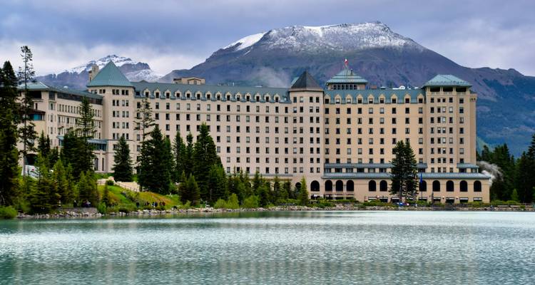 Grand hotel aan een turquoise meer met besneeuwde bergen op de achtergrond.