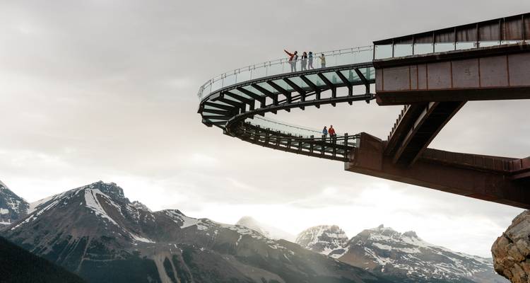 Een groep mensen op een gebogen glazen loopbrug met uitzicht op bergen.