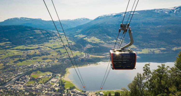 A modern gondola cabin descends toward a lakeside town framed by expansive mountain landscapes and a shimmering lake far below.