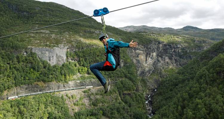 A solo rider waves mid-air on a zipline stretching across a deep gorge with rugged cliffs and a forested valley below.