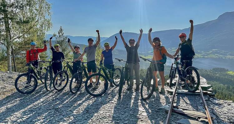 A jubilant group of mountain bikers raises their arms in celebration on a ridge overlooking a fjord and mountains.