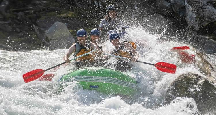 A raft packed with paddlers crashes through roaring white-water rapids, water spraying dramatically all around.