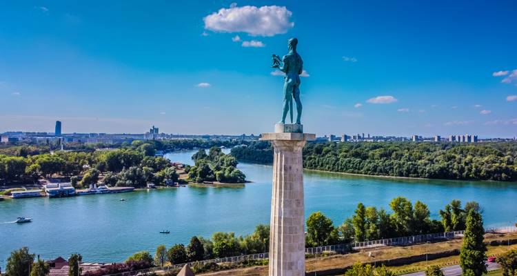 Grande statue surplombant une rivière et un paysage urbain sous un ciel bleu.