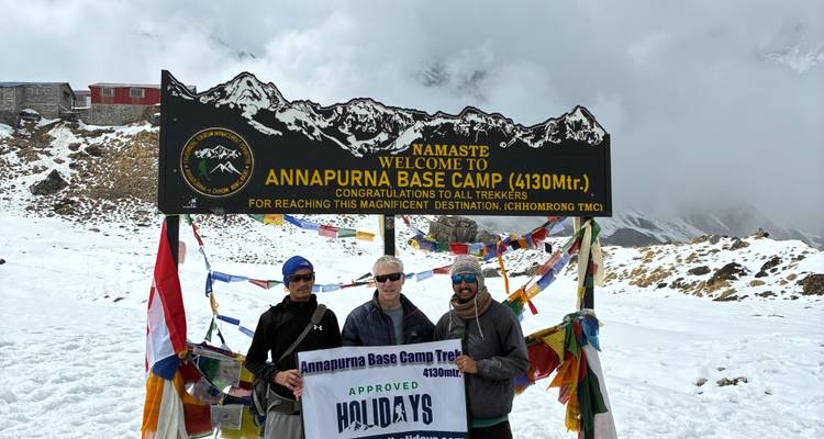 Des trekkeurs posant devant un panneau coloré recouvert de drapeaux de prière au camp de base de l'Annapurna dans les montagnes enneigées.
