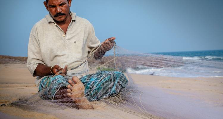 A man sitting on a beach mending a fishing net with the sea in the background.
