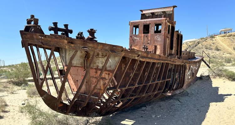 Een roestend scheepswrak rust op woestijnzand onder een heldere blauwe lucht.