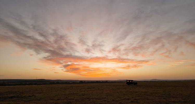 Vurig rood zonsonderganglucht boven open savanne met silhouet van een safariauto aan de horizon.