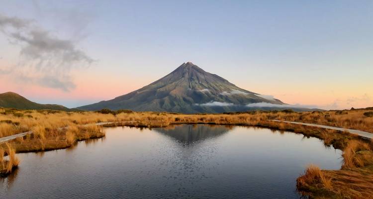 Zonsondergangzicht van Mount Taranaki weerspiegeld in een vijver, omgeven door droog gras.