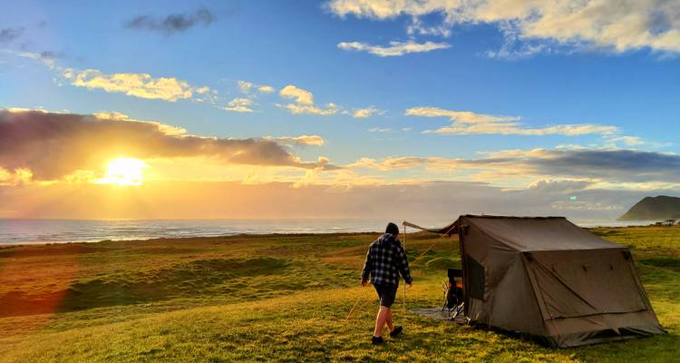 Tentopbouw op een heuvel met uitzicht op zee tijdens zonsondergang, met een persoon die er naartoe loopt.