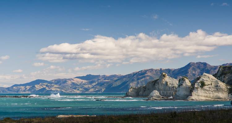 Paysage côtier rocheux avec des vagues et des montagnes au loin.