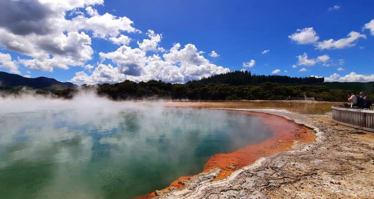 Hot spring with steam and colorful mineral deposits surrounded by lush greenery under a cloudy sky.