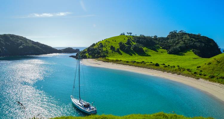 Turquoise bay with sailboat anchored beside lush green peninsula under bright sun.