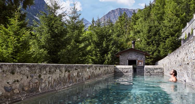 Une personne se relaxant dans un bassin de source chaude entourée d'une forêt avec des montagnes en arrière-plan.